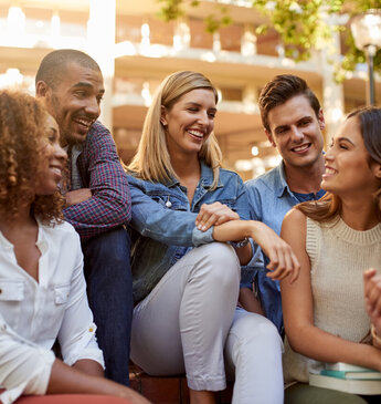 Gruppe fröhlicher junger Erwachsener verschiedener Ethnien, die gemeinsam auf einer Außentreppe sitzen und sich lebhaft unterhalten, symbolisch für Freundschaft und Diversität.