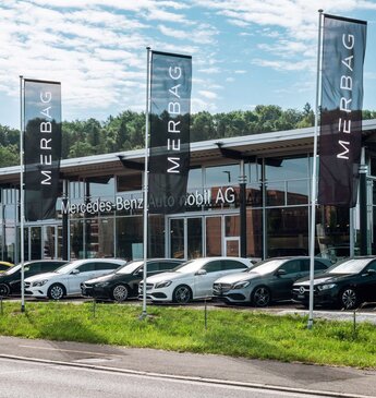 Eine Reihe von neuen Mercedes-Benz Fahrzeugen vor der modernen Glasfassade der Merbag Filiale in Winterthur, umgeben von grüner Landschaft.