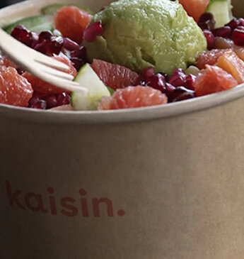 Eine Hand hält eine Gabel über eine appetitlich angerichtete Poké Bowl von kaisin., gefüllt mit sorgfältig ausgewählten Zutaten wie frischem Fisch, grünen Bohnen, reifen Avocadostücken und Granatapfelkernen, serviert in einer umweltfreundlichen Pappschale.